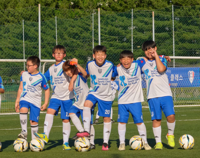 축구에 재능 있는 다문화청소년으로 구성된 유소년클럽 입단 지원반 아이들이 운동장에서 환하게 웃고 있습니다.